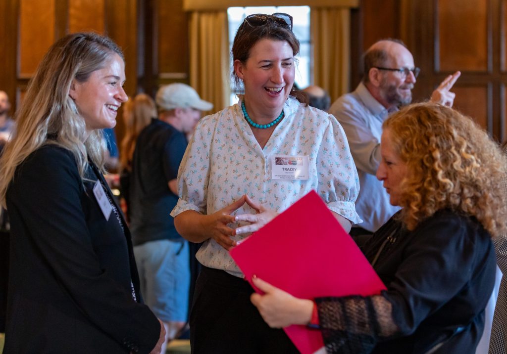 Two women smiling at a third woman opening up a red folder.