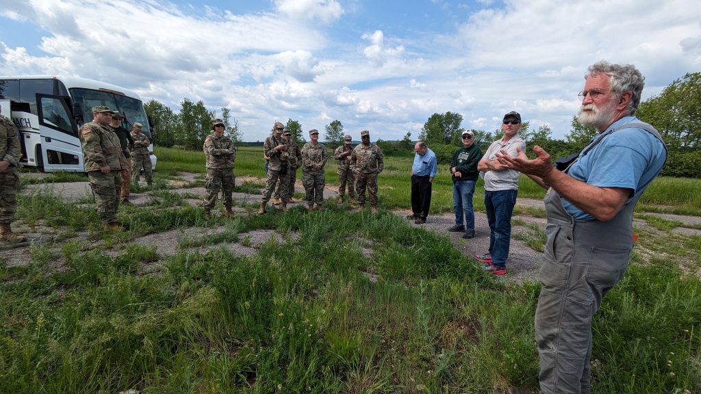 A group of soldiers in camouflage uniforms alongside three other men in casual clothing listen attentively to an older man with a gray beard in overalls, speaking outdoors by a parked bus.